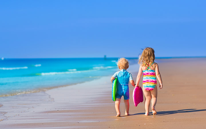 Crianças caminhando na orla da praia segurando uma prancha Crianças caminhando na orla da praia segurando uma prancha